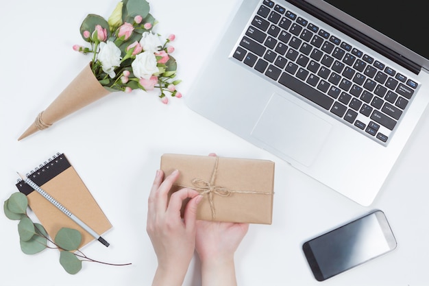 Top view of workspace with laptop, woman hands with present box, notebook, mobile phone