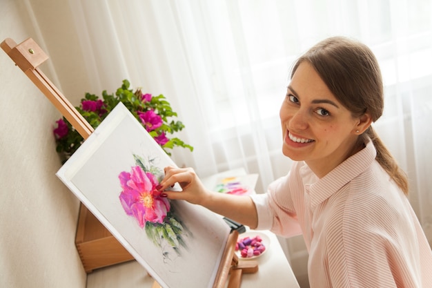 Top view of young caucasian pretty girl artist paints sketch of pink flower sitting at table with bouquet of rose and peonies with painting supplies and dry pastels. Creativity and hobby concept