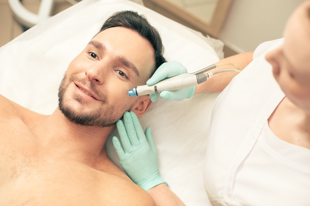 Top view of a young man expressing joy while lying on the pillow and cosmetologist nourishing his skin on the cheek with a help of a modern device