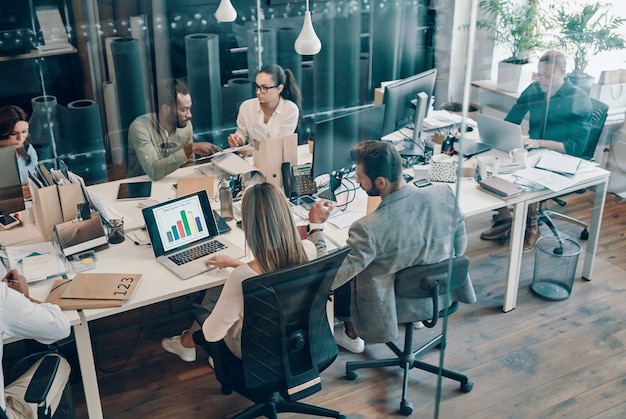 Top view of young modern people in smart casual wear communicating and using modern technologies while working in the office