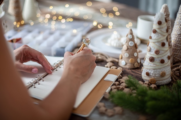 Top view of young woman is writing goals for New Year 2021 on diary on desk with handcrafted Christmas Tree decorations, stars, buttons and tea with marshmallow. Concept of planning new goals.