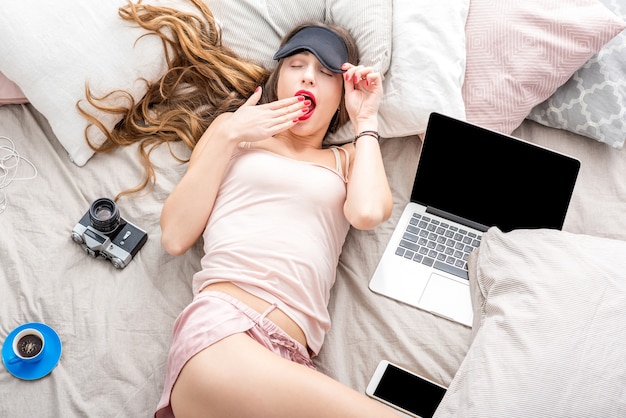 Top view on the young woman lying with eyesight on the bed with laptop, phone and photo camera. Resting after the work