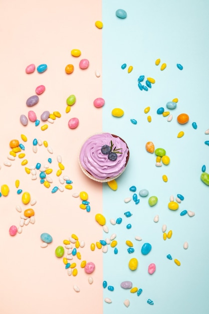 Top view of yummy cupcake with cream and blueberries placed amidst colorful sweets on peach and blue background