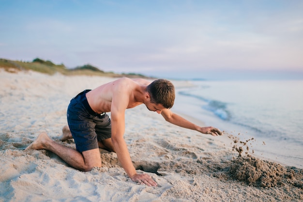 topless man in shorts standing on knees and digging pit by hands on beach beyond sea in summer evening.