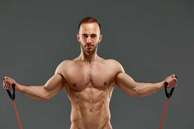 A topless sportsman performs fitness exercises with rubber band in a studio on gray background Bodybuilding Athlete