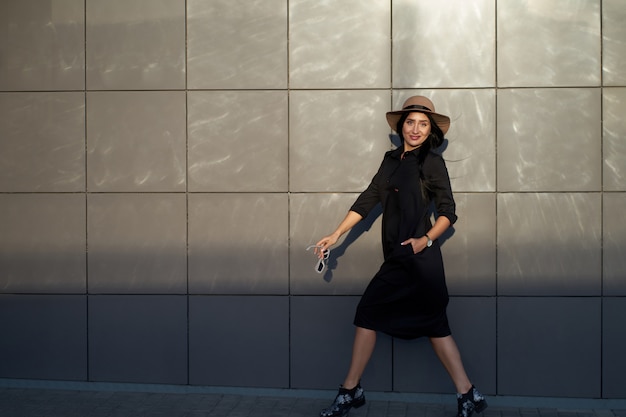 Total look for every day. Cheerful young active attractive brunette girl wearing stylish black dress and fashionable hat. Model in trendy youth outfit walking on urban wall background.