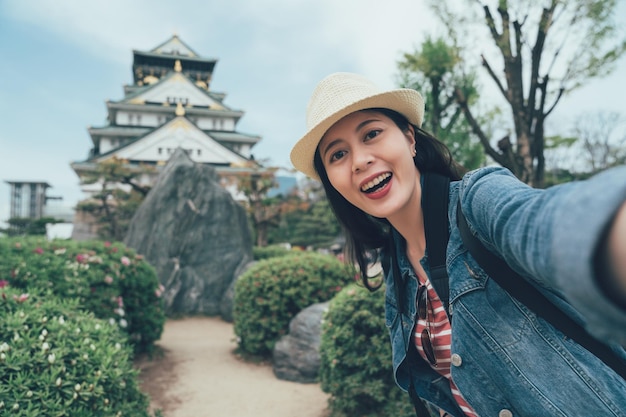 Tourism in japan. happy female tourist take photo selfie in front of osaka castle. asian woman backpacker in straw hat standing in japanese style garden with big rock make self portrait with palace.