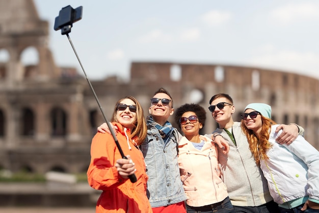 tourism, travel, people, leisure and technology concept - group of smiling teenage friends taking selfie with smartphone and monopod over coliseum ruins in rome background