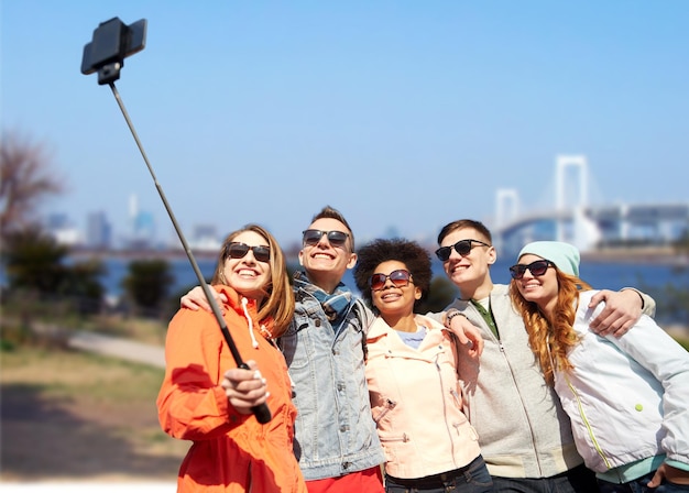 tourism, travel, people, leisure and technology concept - group of smiling teenage friends taking selfie with smartphone and monopod over rainbow bridge in tokyo city