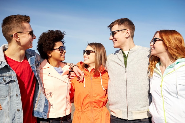 tourism, travel, people, leisure and teenage concept - group of happy friends in sunglasses hugging and talking on city street