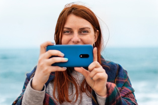 Tourist caucasian red hair girl takes pictures or recording video on smartphone on the beach near the ocean