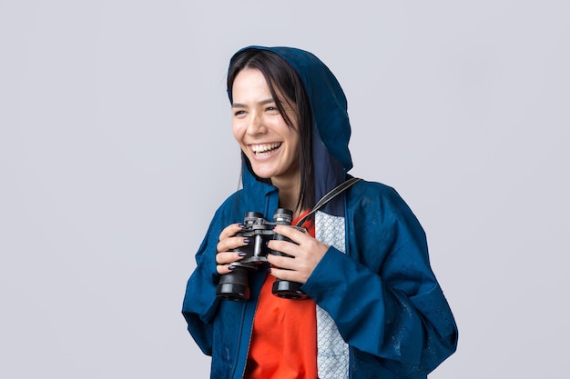 A tourist girl in a blue raincoat holds binoculars in her hands and looks into the distance, spies.