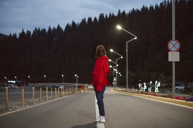 Tourist girl in a red raincoat strolling near the mountain fir forest in the street lights