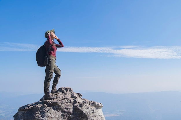 Tourist girl with backpack standing on the stone at the mountain, wear hat and looking panoramic view at Pha Mak Dook Phukradung National park at Thailand.