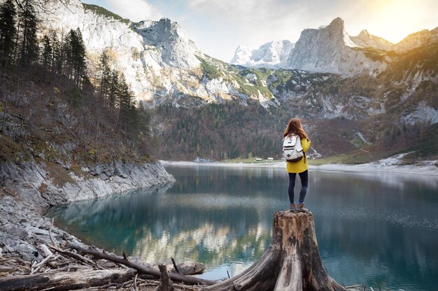 tourist girl with a backpack stands on a huge stump on the shore of a mountain lake