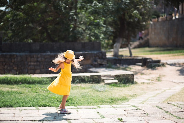 A tourist girl in a yellow hat and sundress dancing along the street of the old town made of stone with a fortress Sightseeing tour