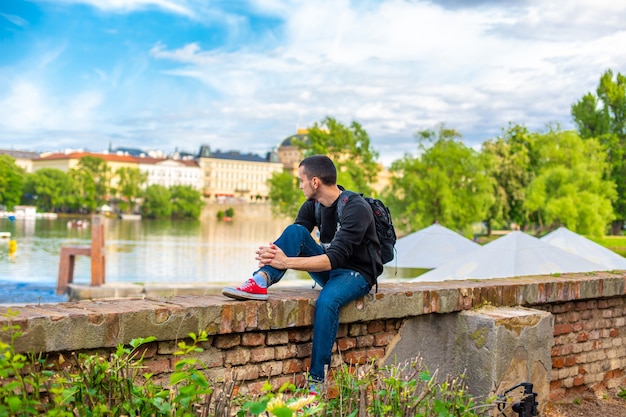 A tourist guy with a backpack sits on the embankment in Prague overlooking the Vltava river.