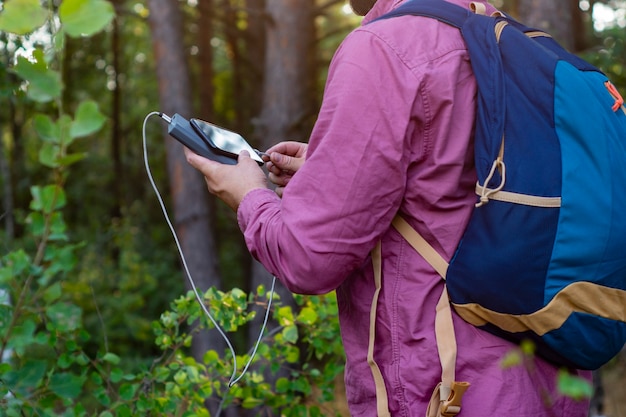 Tourist holds a smartphone with a portable charger in his hands. Man with a Power Bank charges the phone against the backdrop of nature and forest.