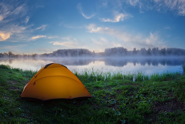 Tourist orange tent on green grass of foggy lake at sunrise