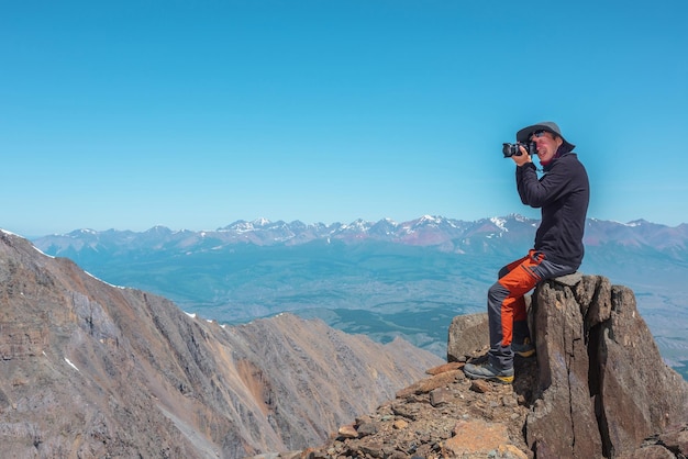 Tourist sitting on stone near abyss edge on high altitude under blue sky in sunny day Man with camera on high rock near precipice edge with wonderful view from above to large mountain range in away