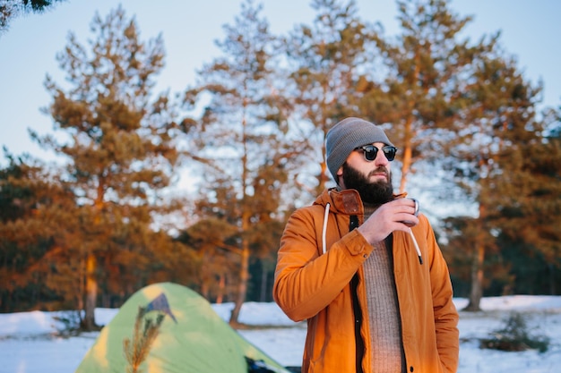 A tourist standing near a tent with a cup of tea or coffee in the warm rays of the setting sun in a winter forest