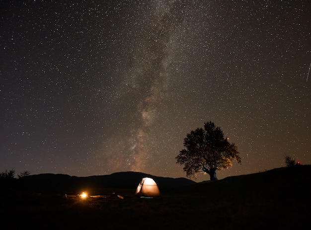 Tourist tent and burning campfire on green valley under dark starry sky.