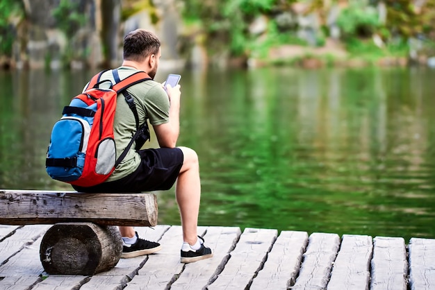 A tourist with a backpack on his back sits on a tree bench by the lake and looks at the phone