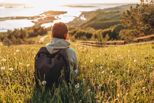 Tourist with backpack sitting on top of hill in grass field and enjoying beautiful landscape view Rear view of teenage boy hiker resting in nature Active lifestyle Concept of local travel