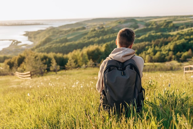 Tourist with backpack sitting on top of hill in grass field and enjoying beautiful landscape view Rear view of teenage boy hiker resting in nature Active lifestyle Concept of local travel