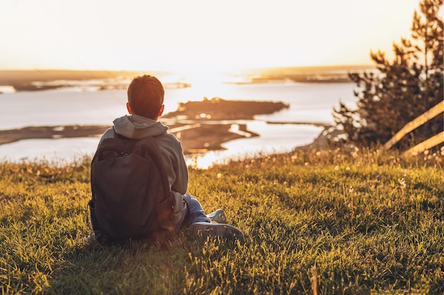 Tourist with backpack sitting on top of hill in grass field and enjoying beautiful landscape view Rear view of teenage boy hiker resting in nature Active lifestyle Concept of local travel