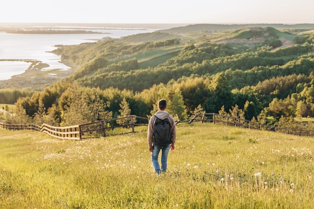 Tourist with backpack standing on top of hill in grass field and enjoying beautiful landscape view Rear view of teenage boy hiker resting in nature Active lifestyle Concept of local travel