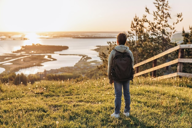 Tourist with backpack standing on top of hill in grass field and enjoying beautiful landscape view Rear view of teenage boy hiker resting in nature Active lifestyle Concept of local travel