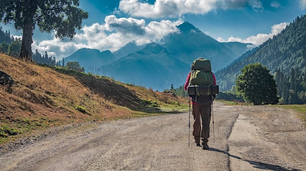 Tourist with backpack and trekking poles on road at mountains background at sunny day