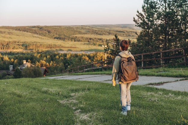 Tourist with backpack walking on top of hill in grass field and enjoying beautiful landscape view Rear view of teenage boy hiker resting in nature Active lifestyle Concept of local travel