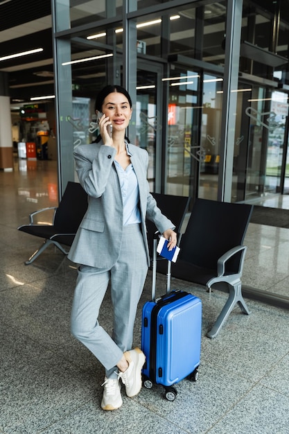 Tourist with blue suitcase is talking on phone while waiting for a hotel transfer in an airport lounge Young attractive woman standing in waiting room of airport and talking on phone