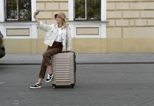 Tourist woman in a European city, tourism in Europe. a young girl dressed in a Panama hat sits on a large suitcase makes a selfie on her phone. Copy space.