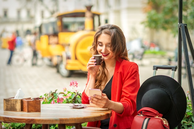 Tourist young caucasian woman in red jacket with coffee cup at the table in cafe outdoors