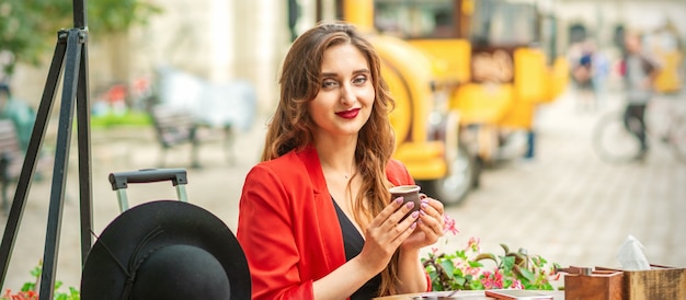 Tourist young caucasian woman in red jacket with coffee cup at the table in cafe outdoors.