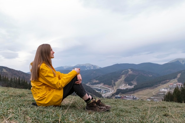 Tourist young woman sits on hillside with cup in autumn Female traveler looks at the mountains