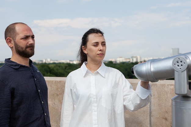 Tourists couple spending relationship anniversary on building terrace enjoying summer holiday. Woman looking through telescope at metropolitan city admiring beautiful landscape from observation point