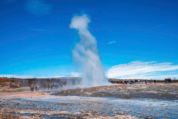 Tourists enjoying view of strokkur geyser eruption in valley against blue sky