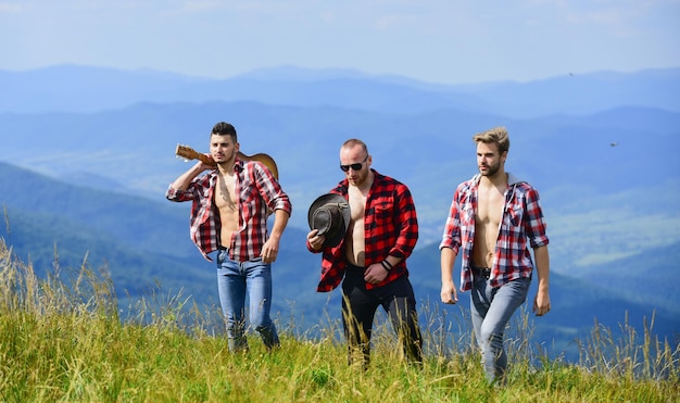 Tourists hiking concept Hiking with friends Friendly guys with guitar hiking on sunny day Enjoying freedom together Group of young people in checkered shirts walking together on top of mountain