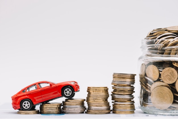 Toy car going up on the increasing stack of coins near the coins jar against white background