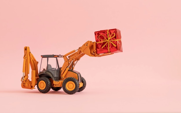 Photo toy excavator carries a gift box tied with a bow. pink background. copy space,