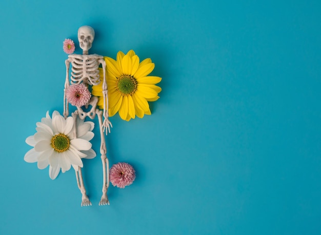 Toy skeleton in white and yellow chrysanthemum flowers on blue background, creative Concept Halloween, the idea of life and death, free space for text