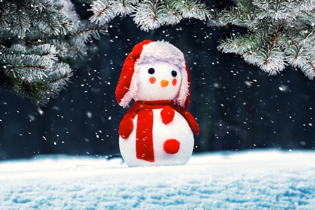 Toy snowman in the snow near a snowy spruce during a snowfall, New Year and Christmas greeting card