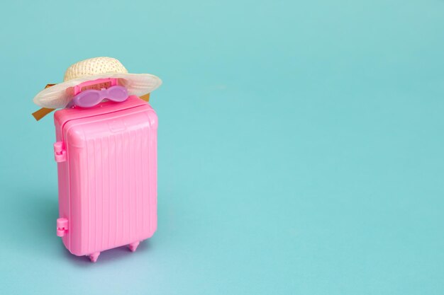 Toy suitcase with sunglasses and a hat on a light turquoise background with copy space travel content