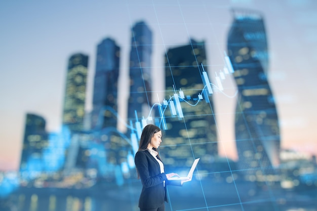 Trade finance and market concept Attractive young caucasian businesswoman using laptop on blurry city background with forex chart Double exposure