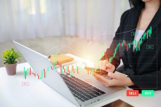 Trader or businesswoman hands showing virtual hologram price graph and indicator, red and green candlestick chart stock trading on laptop computer and smartphone. Investing on stock concept.