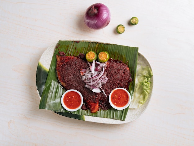 Traditional BBQ stingray served on banana leaf with onions red chillies sauce and lemon on a white plate top view